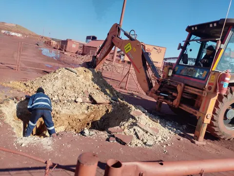 A worker in a trench by a TLB, digging out a pipe line on a red-soil site