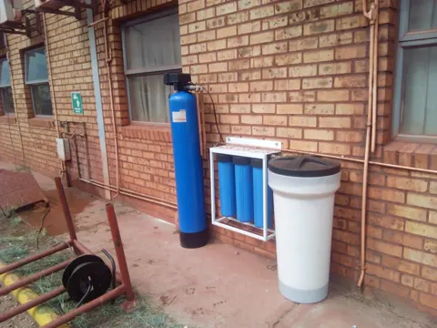 A blue water filter tank, a three-cartridge filter and a white brine tank fitted against a face-brick wall