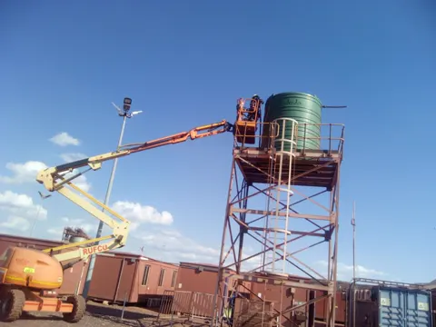 A green water tank on a tall steel tower, with a cherry picker lifting a worker up to it
